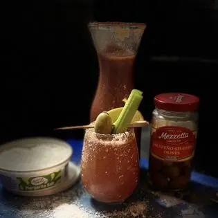 Bloody Mary with a celery stalk and olive next to a jar of jalapeno stuffed olives